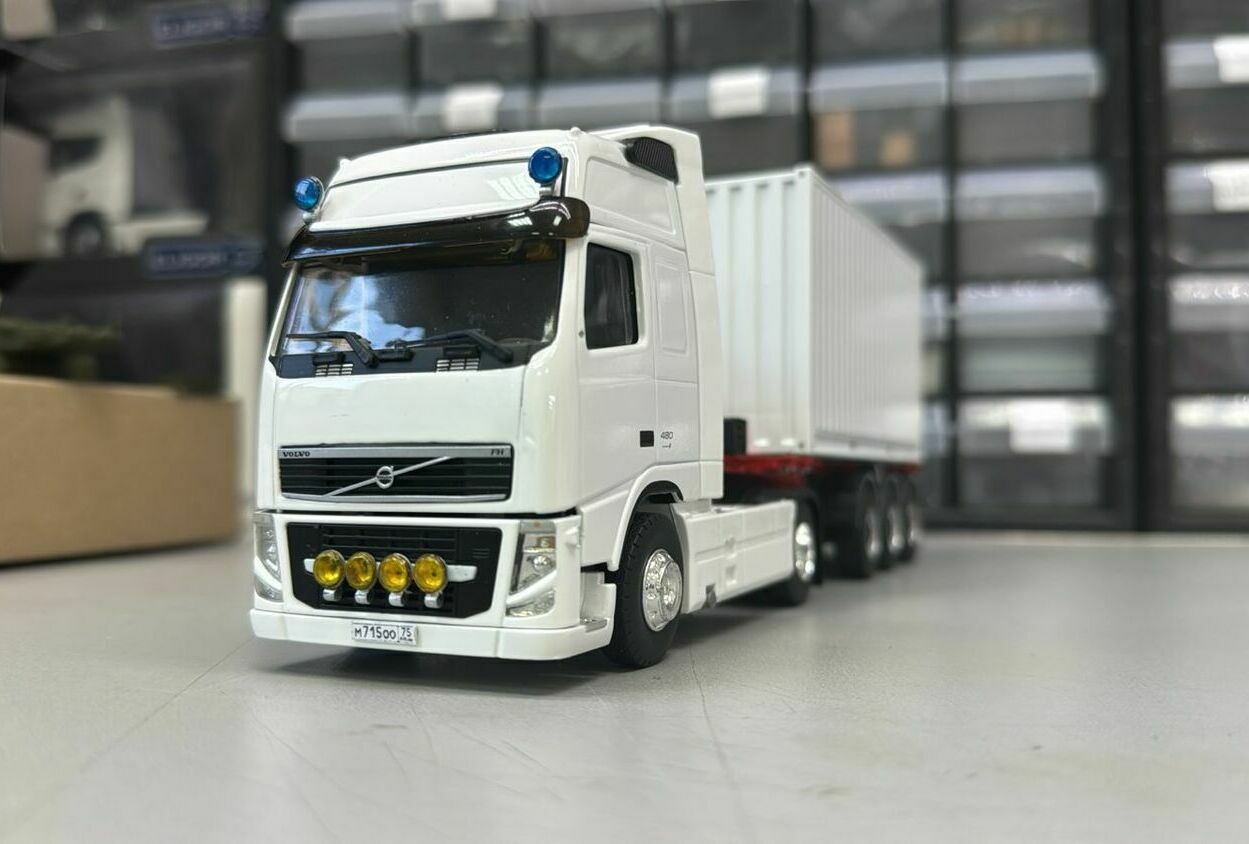 Тягач компоновки с высокой спальной кабиной (high-roof sleeper cab).   • Передняя светотехника: основные фары, блок дополнительных противотуманных/дальнобойных фар на переднем бампере.   • Солнечный козырёк над ветровым стеклом, сигнальные огни на крыше кабины.   • Фирменная решётка радиатора Volvo с логотипом и обозначением модели FH и мощности «480».   • Смоделированные элементы ходовой части: односкатные шины на управляемой передней оси и ведущей задней.   • Сцепка с трёхосным полуприцепом прямоугольного сечения (контейнер/фургон).    Модель установлена на ровной поверхности, на фоне стеллажей с ячейками для хранения и коробок с другими моделями.