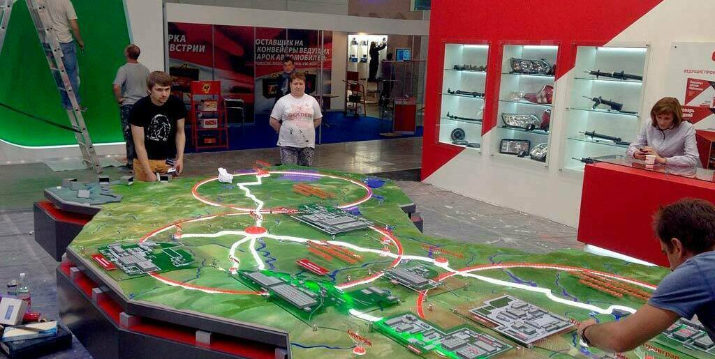 3D масштабный макет территории промышленного предприятия с детализированными моделями зданий, имитацией ландшафта и системой светящихся маршрутов логистики. Интерактивная инсталляция на выставочном стенде, демонстрирующая структуру производственного кластера в сфере автопрома. На макете видны рельеф местности, водоемы, подъездные пути и зоны цехов, подсвеченные для наглядности технологических цепочек.