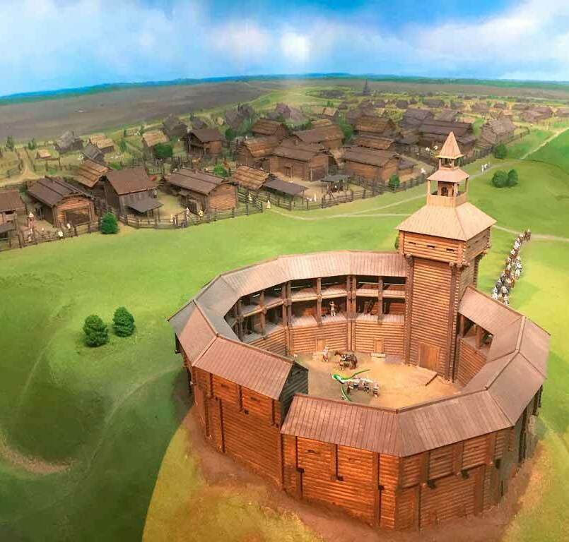 Макет изготовленный для музея на заказ На макете изображён небольшой деревянный острог, раскинувшийся среди волнистых зелёных холмов. Впереди, почти вплотную к зрителю, высится сторожевая башня с открытой дозорной площадкой под остроконечной крышей: часовой, укрывшись в тени навеса, наблюдает за дорогой. За башней виден целый посёлок из срубленных из тёса изб и амбаров, окружённых частоколом; между домами тянутся узкие улицы, дворики и загоны для скота. От дальних пределов макета к воротам крепости тянется цепочка всадников: вооружённая дружина в кольчугах и шлемах, выстроенная узкой колонной. Их кони ступают по утоптанной дороге, режущей сочную зелень холмов. За высоким небесным сводом, расписанным мягкими облаками, простирается степь, уходящая к горизонту. Всё вместе создаёт образ тихого, но настороженного пограничного мира, живущего под защитой деревянных стен.