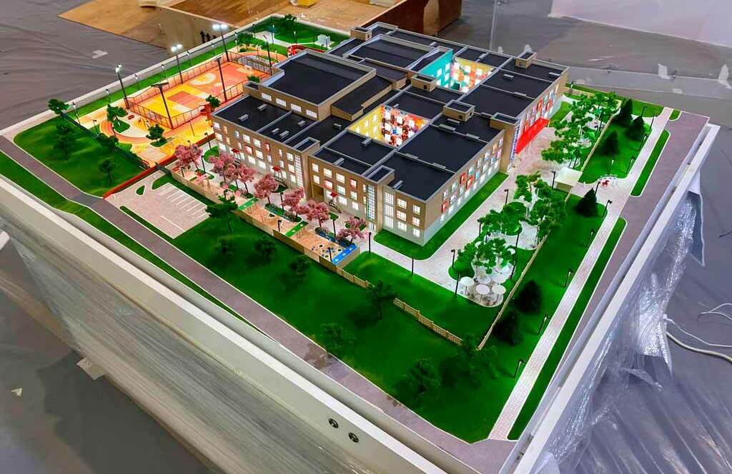 Детализированный архитектурный макет школьного комплекса с благоустроенной территорией: трёхэтажное здание школы, спортивная и детская площадки, зелёные зоны, аллеи и парковочная зона — визуализация образовательного объекта для презентации проекта Масштабный архитектурный макет современного школьного комплекса: трёхэтажное здание с освещёнными окнами, чётко выделенными входами и архитектурными деталями фасада. На прилегающей территории — огороженная спортивная площадка с разметкой для игр, детская игровая зона, благоустроенные аллеи с миниатюрными деревьями и кустарниками, зелёные газоны. Присутствуют парковочная зона, подъездные пути, уличные фонари, декоративные элементы ландшафта (беседки, клумбы). Макет демонстрирует гармоничное сочетание образовательной инфраструктуры и элементов ландшафтного дизайна, позволяет оценить планировку комплекса, распределение зон отдыха и активности, а также логистику передвижения вокруг здания. Подходит для презентации инвестиционного или градостроительного проекта.