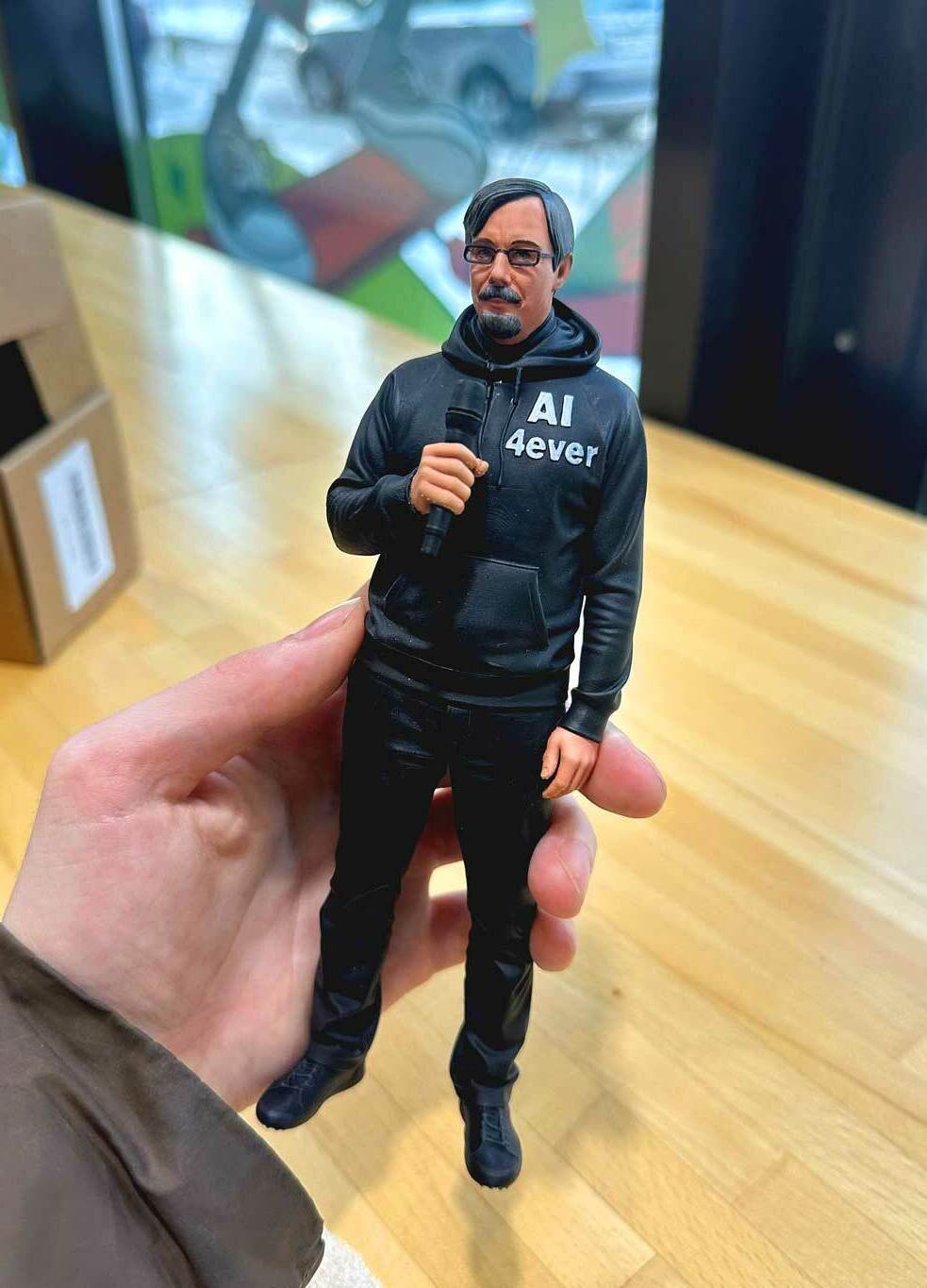 Реалистичная 3D-фигурка человека на заказ по фото: детализированная статуэтка с микрофоном, напечатанная на 3D-принтере. Кастомная модель в чёрной худи с надписью — идеальный сувенир для коллекционеров и поклонников публичных выступлений! 3D-фигурка мужчины, изготовленная по фотографии на 3D-принтере. Статуэтка изображает человека в расслабленной позе с микрофоном в руке. Одет в чёрную худи с надписью «AI ever», чёрные брюки, носит очки и аккуратную бороду. Высокая детализация: проработаны черты лица, текстура одежды, поза передаёт атмосферу публичного выступления. Фигурка отлично подойдёт для украшения офиса, в качестве оригинального подарка или элемента коллекции 3D-моделей. Пример персонализированной печати с сохранением индивидуальных особенностей.