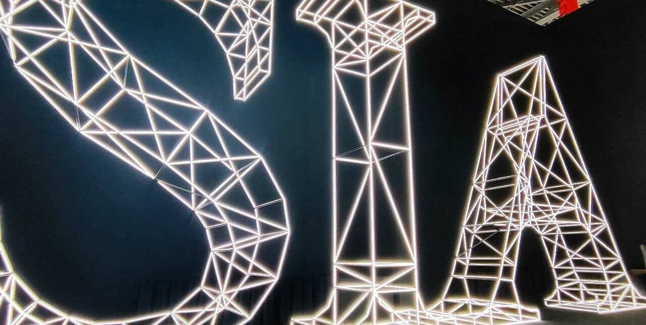 3‑D масштабный макет объёмных букв SIA: белая полигональная сетка, яркая светодиодная подсветка, чёткие грани и каркасная структура; индустриальный стиль, тёмный фон, зона отдыха с мебелью, акцент на современном оформлении выставочных и коммерческих площадок3‑D масштабный макет букв SIA: полигональная каркасная структура с равномерной светодиодной подсветкой, белый контур, чёткая геометрия граней; композиция размещена в тёмном пространстве с индустриальным потолком, справа — зона с креслами, слева — экран.