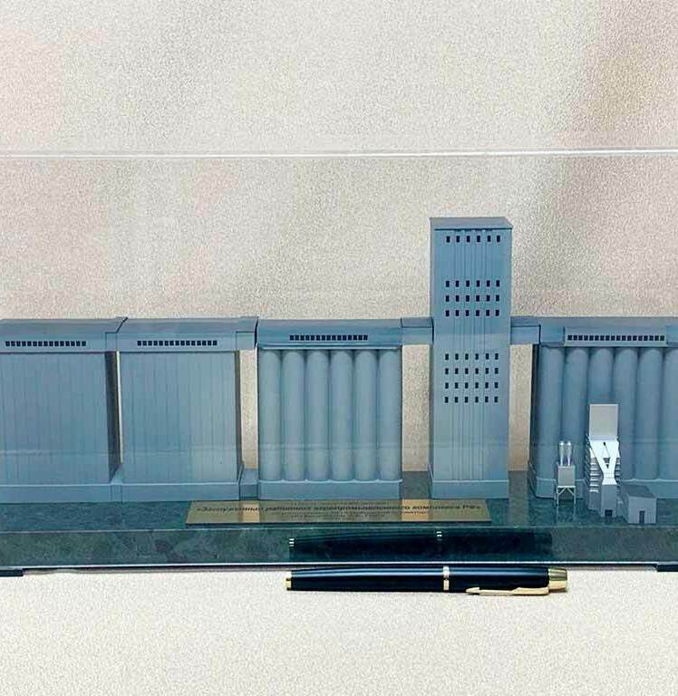 На макете видно  несколько секций сооружений серого цвета на общем основании. • Слева — прямоугольные блоки, похожие на складские или технологические корпуса. • По центру и справа — ряды вертикальных цилиндрических силосов, соединённых верхними галереями. • В центре — высокая прямоугольная башня с рядами небольших проёмов (головная башня элеватора). • Справа у силосов — мелкие детали: лестницы, площадки, оборудование. • Внизу на основании — металлическая табличка с названием, перед макетом лежит ручка для масштаба. В целом это аккуратная масштабная модель зернохранилища/элеваторного комплекса: силосы, перегрузочные галереи и технологическая башня.