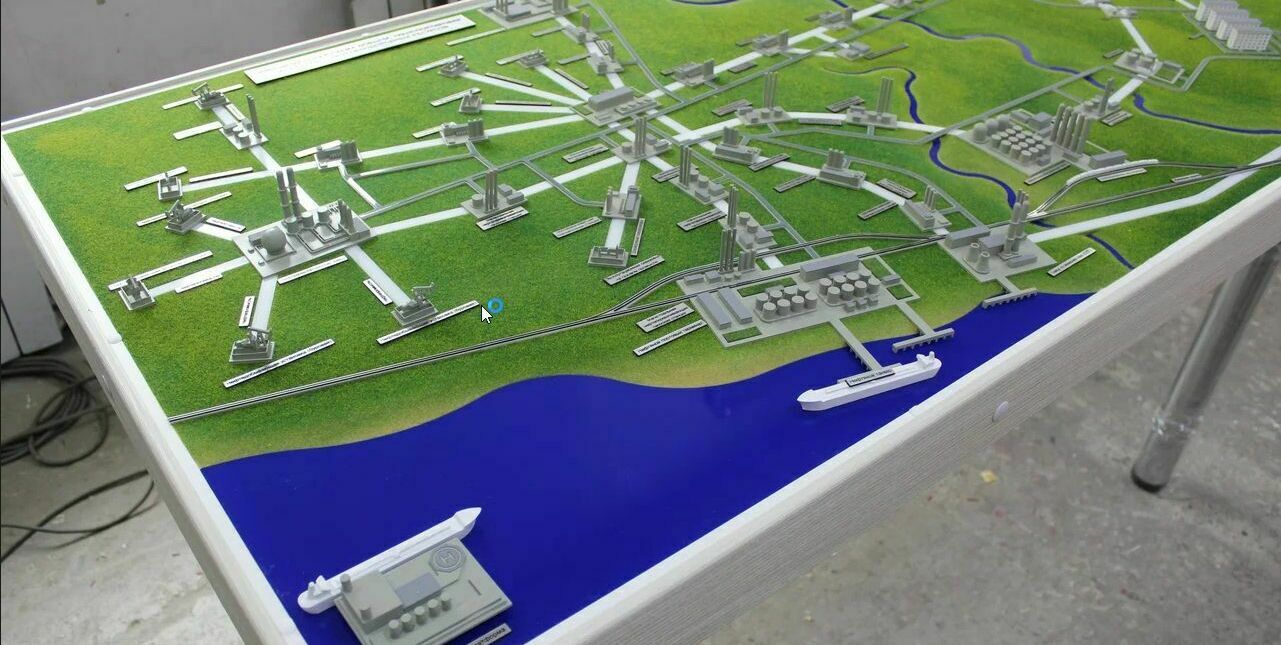 Масштабный макет-схема городской инфраструктуры, изготовленный на заказ в Макетной мастерской: детализированное отображение зданий, дорог, водного объекта и транспортных средств — инструмент для презентации градостроительных проектов Детальный макет-карта архитектурной направленности, демонстрирующий городскую инфраструктуру: множество серых зданий и сооружений, соединённых сетью дорог и путей. Зелёный фон символизирует травяное покрытие или растительность, синяя область в нижней части макета изображает водоём или реку. На макете размещены модели кораблей и других транспортных средств, что позволяет наглядно представить логистику и взаимодействие различных элементов городской среды. Идеален для образовательных целей, презентаций проектов и музейных экспозиций.