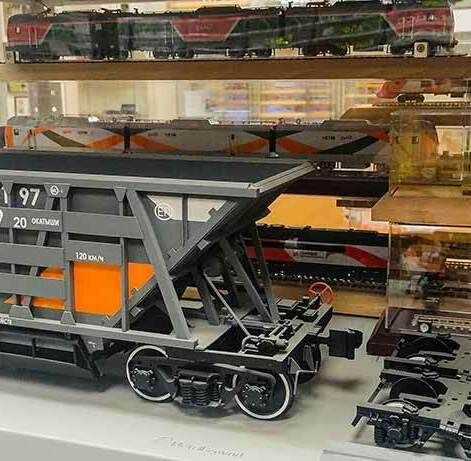 Изготовление детальных масштабных моделей железнодорожной техники: локомотивы, грузовые вагоны-хопперы и подвижной состав на заказ Коллекционный макет грузового вагона-хоппера для перевозки окатышей с высокой детализацией на фоне стендовых моделей локомотивов и поездов РЖД в масштабе