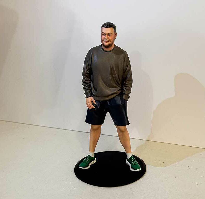 Реалистичная 3D-фигурка человека по фото на заказ — детализированная статуэтка в повседневной одежде, напечатанная на 3D-принтере с высокой точностью. Уникальный сувенир с портретным сходством! 3D-фигурка мужчины, изготовленная по фотографии на 3D-принтере. Фигура в расслабленной позе: одна рука в кармане, другая свободно опущена. Одет в тёмно-серый свитшот, чёрные шорты и зелёные кроссовки. Статуэтка установлена на круглой чёрной подставке. Высокая детализация: проработаны черты лица, текстура одежды, пропорции тела. Пример персонализированной 3D-печати с передачей индивидуального стиля и характера. Подходит для подарка, коллекционирования или декора.