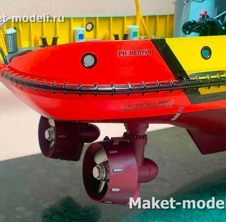 Это фото детализированного стендового макета корпуса буксира. Видна носовая/скуловая часть с массивным привальным брусом, иллюминаторами и маркировкой «Осторожно винт / Take care side propeller». Под корпусом смонтированы два кольцевых движителя: винты в кольцевых насадках (тип Корта) на поворотных колонках, работающих как основные азимутальные пропульсоры/подруливающие устройства. На борту нанесено название «РН Нептун».