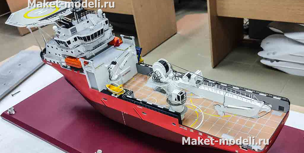 3‑D масштабная модель судна обеспечения: красный корпус, белая надстройка, детализированные винторулевые колонки и палубное оборудование»Перед нами 3‑D масштабная модель судна обеспечения — высокодетализированная копия с красным корпусом, белой надстройкой, палубными кранами, спасательными шлюпками и винтами; экспонат размещён на опорах в выставочном пространстве.