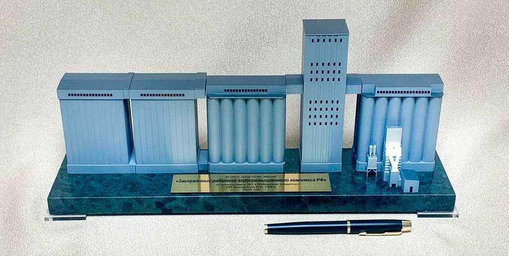 Несколько корпусов хранения: блоки цилиндрических силосов (силосные батареи) и прямоугольные секции складов. • Центральная рабочая (норийная) башня с окнами — узел вертикального транспортирования зерна (нории, распределение по силосам). • Надсилосные и межкорпусные галереи — закрытые конвейерные переходы между секциями. • Узлы приёма/подготовки зерна у правого блока (оборудование очистки/аспирации, загрузочные устройства). • Основание‑подставка с табличкой (памятный/наградной макет для работников АПК).  Назначение объекта: приём, транспортирование, очистка и распределение зерна по силосам для длительного хранения и последующей отгрузки.