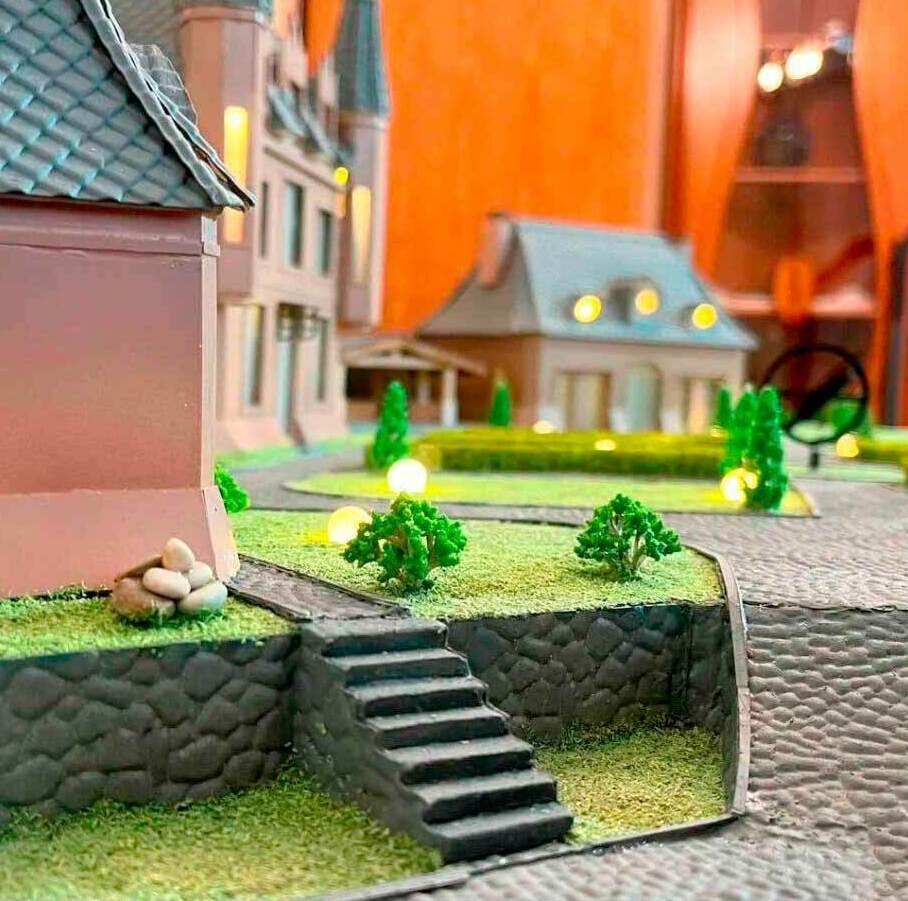 Масштабная модель усадебной застройки с несколькими зданиями, выполненными в классическом стиле с элементами барокко. На переднем плане — участок с каменной лестницей, ведущей к дому, и подпорной стенкой из имитации каменной кладки. Территория оформлена миниатюрными кустарниками и газоном, создающими ощущение ухоженного сада. В центре композиции — дом с высокой башней и черепичной крышей, рядом расположен двухэтажный особняк с мансардой и большими окнами. Между зданиями проложены мощёные дорожки, установлены декоративные фонари с тёплым светом. Модель демонстрирует гармоничное сочетание архитектуры и ландшафтного дизайна, включает детализированные элементы благоустройства: клумбы, деревья, мощение. Подходит для визуализации проектов частной застройки, презентаций перед инвесторами и использования в экспозициях архитектурных бюро.