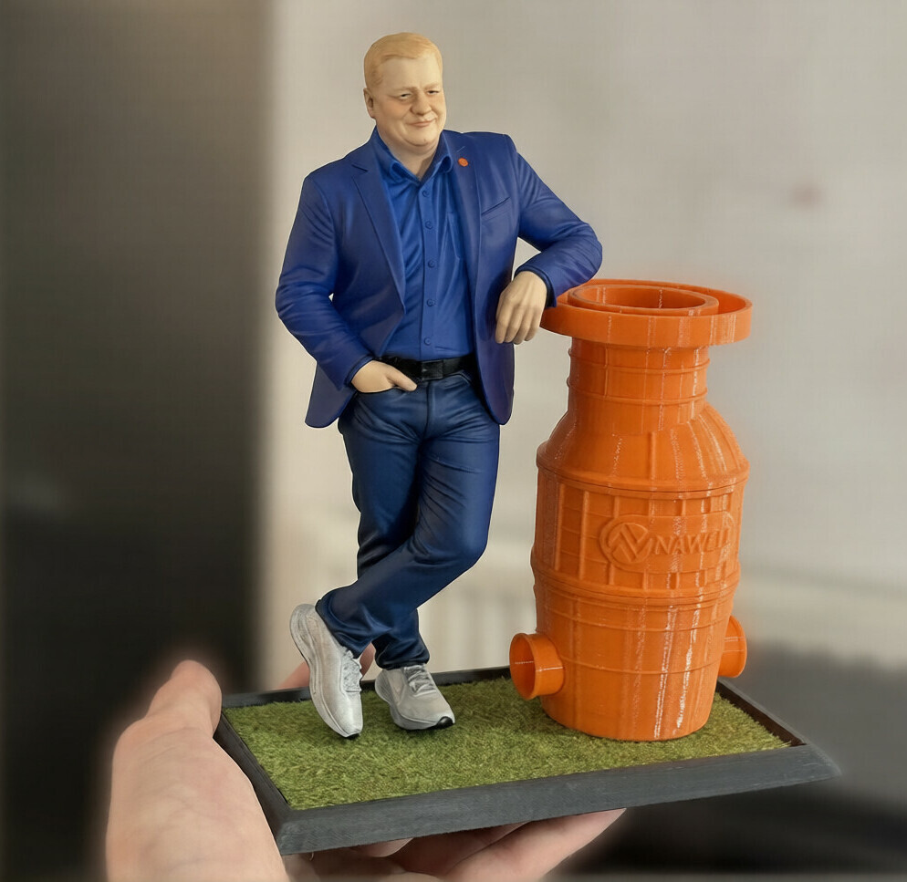 3D-фигурка человека по фото на заказ: реалистичная статуэтка в деловом костюме с оранжевым промышленным оборудованием — печать на 3D-принтере с детализацией одежды и позы Портретная 3D-фигурка мужчины, изготовленная по фотографии на 3D-принтере. Фигура в расслабленной позе: одна рука опирается на ярко-оранжевое промышленное оборудование с логотипом «WINAWELL», другая — в кармане. Мужчина одет в синий костюм, голубую рубашку и белые кроссовки. Статуэтка установлена на прямоугольной подставке с имитацией травяного покрытия. Пример персонализированной 3D-печати с высокой детализацией: проработаны черты лица, текстура одежды, поза и аксессуары. Подходит для корпоративного подарка, декора офиса или коллекционирования.
