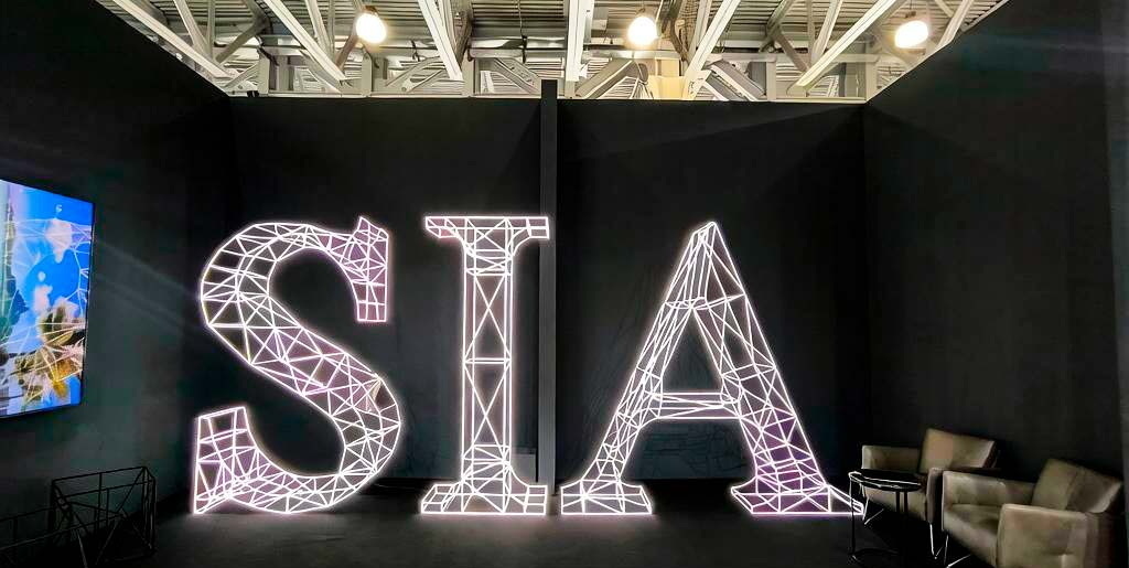 3‑D масштабный макет объёмных букв SIA: белая полигональная сетка, яркая светодиодная подсветка, чёткие грани и каркасная структура; индустриальный стиль, тёмный фон, зона отдыха с мебелью, акцент на современном оформлении выставочных и коммерческих площадок3‑D масштабный макет букв SIA: полигональная каркасная структура с равномерной светодиодной подсветкой, белый контур, чёткая геометрия граней; композиция размещена в тёмном пространстве с индустриальным потолком, справа — зона с креслами, слева — экран.