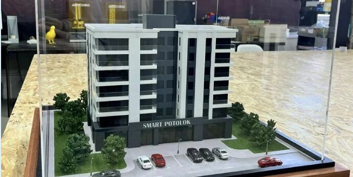 3D-масштабный макет современного многоэтажного жилого здания с подсветкой фасадов. Модель демонстрирует планировку комплекса: главный корпус с балконами и большими окнами, входная группа, крытая парковка с несколькими автомобилями, благоустроенные зелёные зоны с деревьями и кустарниками по периметру. Видны элементы городской инфраструктуры — проезжая часть с разметкой, тротуар, уличные фонари. Макет выполнен с высокой детализацией, включая текстуры фасадов, ландшафт и миниатюрные автомобили, что позволяет наглядно представить будущий объект и его интеграцию в городскую среду. Идеален для презентации девелоперских проектов инвесторам и будущим жильцам.