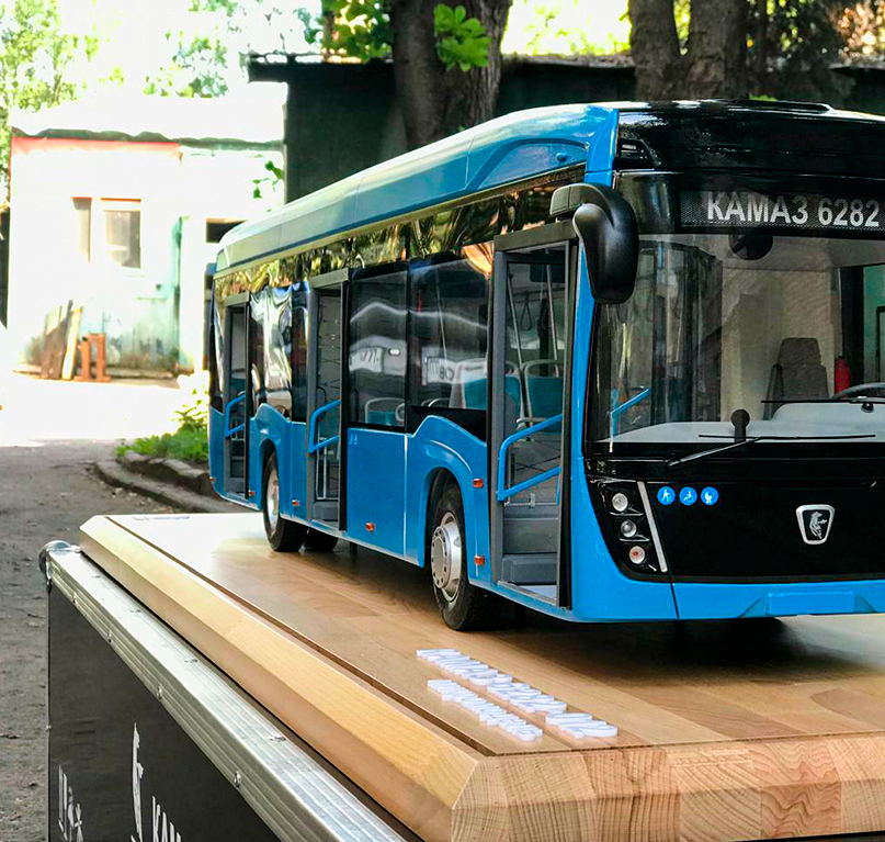 Масштабная модель электробуса КАМАЗ-6282 — детализированная копия городского низкопольного электротранспорта с фирменным логотипом и маршрутным номером, для коллекционеров и презентационных экспозиций, изготовленная на заказ в макетной мастерской Maket-modeli.ru Детализированная масштабная модель электробуса КАМАЗ-6282 в ярком синем цвете с чёрной передней частью. На модели отчётливо видны элементы дизайна: большие панорамные окна, боковые поручни, зеркала заднего вида, светодиодные фары и фирменный логотип КАМАЗ. Модель установлена на деревянной подставке с текстовой вставкой, идеально подходит для выставок, рекламных презентаций и коллекций, посвящённых городскому электротранспорту.