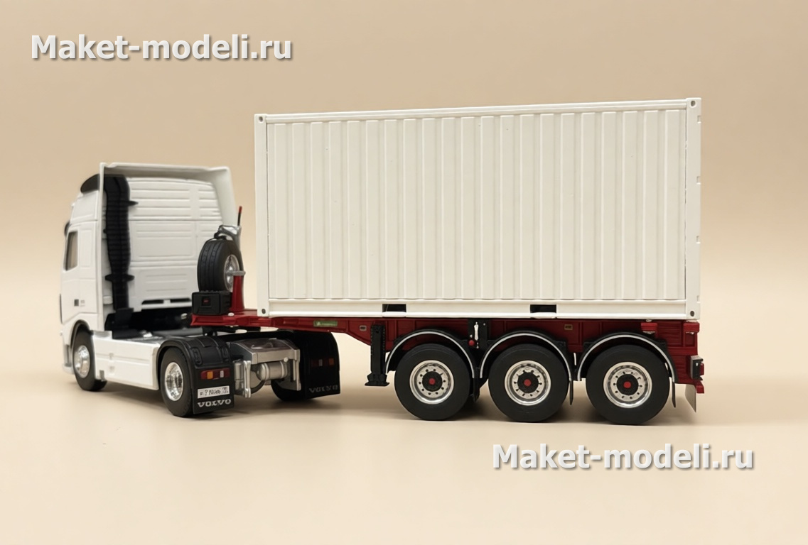Масштабная модель магистрального седельного тягача Volvo FH 480, колесная формула 4×2, с трехосным полуприцепом-фургоном (контейнеровозом) Тягач компоновки с высокой спальной кабиной (high-roof sleeper cab). • Передняя светотехника: основные фары, блок дополнительных противотуманных/дальнобойных фар на переднем бампере. • Солнечный козырёк над ветровым стеклом, сигнальные огни на крыше кабины. • Фирменная решётка радиатора Volvo с логотипом и обозначением модели FH и мощности «480». • Смоделированные элементы ходовой части: односкатные шины на управляемой передней оси и ведущей задней. • Сцепка с трёхосным полуприцепом прямоугольного сечения (контейнер/фургон). Модель установлена на ровной поверхности, на фоне стеллажей с ячейками для хранения и коробок с другими моделями.