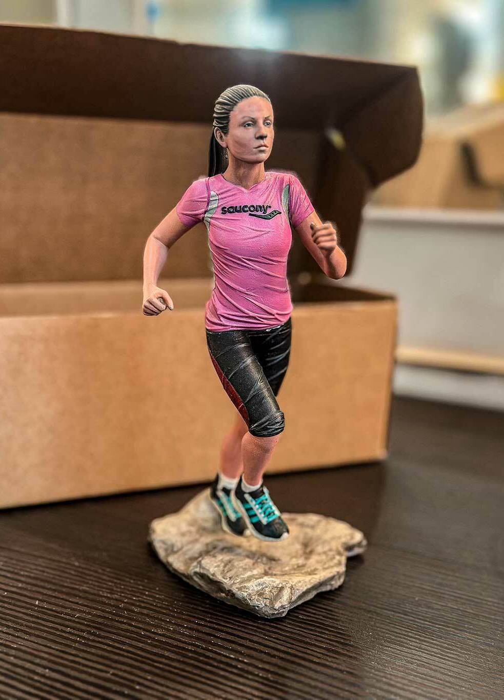 3D-фигурка бегущей девушки, изготовленная на заказ по фото — реалистичная статуэтка на 3D-принтере с детализированной проработкой одежды и позы Статуэтка девушки в спортивной одежде (розовая футболка Saucony, чёрные шорты, кроссовки), запечатлённой в момент бега. Фигурка напечатана на 3D-принтере по фотографии, установлена на основании, имитирующем каменистую поверхность. Пример персонализированной 3D-печати с высокой детализацией: проработаны черты лица, текстура одежды, динамичная поза. Подходит для подарка или коллекционирования.