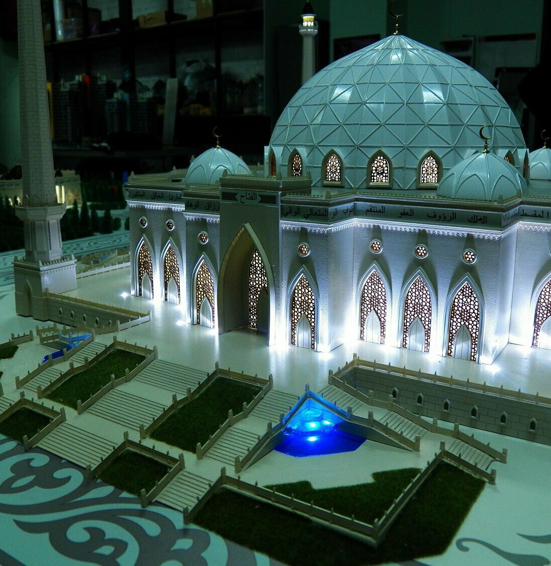 Масштабная модель белой мечети на подиуме: крупный геодезический купол, четыре минарета, ажурные арочные входы с внутренней подсветкой, террасы и ступени, зелёные газоны и синие декоративные бассейны.