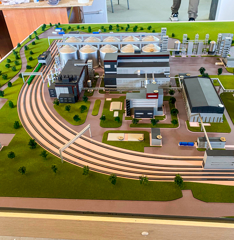 Детализированный макет промышленного предприятия: представлены производственные корпуса, резервуары для хранения сырья, складские помещения, инженерные коммуникации. Особое внимание уделено железнодорожной инфраструктуре — разветвлённая сеть путей с погрузочно-разгрузочными узлами, эстакады. Модель демонстрирует логистику перемещения грузов, взаимосвязь технологических зон, планировку территории с озеленением и подъездными путями. Реалистичная проработка элементов позволяет использовать макет для выставок, образовательных целей, презентации инвестиционных проектов в сфере промышленного строительства.