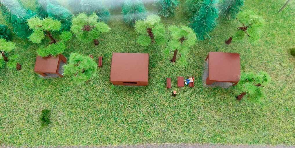  3D-масштабный макет благоустроенной территории коттеджного посёлка: изображены жилые и общественные здания, детская площадка, зелёные насаждения, парковка с автомобилями, огороженные вольеры с животными. Демонстрируется гармоничное сочетание архитектурных объектов и природных элементов — деревья, кустарники, газоны. Видны детализированные элементы инфраструктуры: фонари, дорожки, скамейки. Макет наглядно иллюстрирует планировку участка, распределение зон и взаимодействие построек с окружающим ландшафтом.