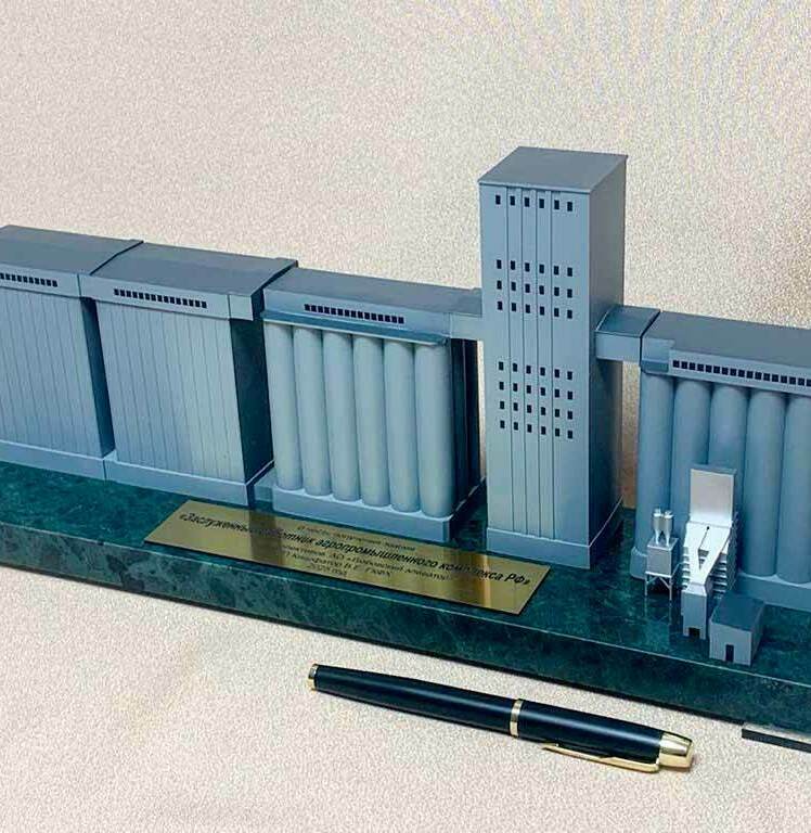 Настольный архитектурный макет промышленного зернохранилища. • Слева — два блока прямоугольных силосов/складских корпусов. • В центре — батарея цилиндрических силосов, соединённых верхними галереями-конвейерами. • Высокая центральная башня — рабочая башня с нориями (узел подъёма и распределения зерна). • Справа — ещё группа цилиндрических силосов и узел приёмки/очистки с загрузочным оборудованием. • Все элементы смонтированы на подставке с табличкой; ручка на переднем плане показывает настольный масштаб модели.  Это демонстрационный макет типового элеваторного (зерноперерабатывающего) комплекса.