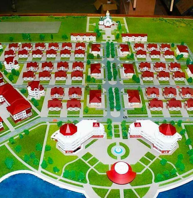 Масштабный макет коттеджного посёлка с инфраструктурой: жилые дома, общественные здания, спортивные площадки и водоём — образец градостроительного проекта для презентации и планирования Детальный макет коттеджного посёлка: упорядоченная сетка жилых домов с красными крышами, окружённых зелёными насаждениями. В центре композиции — общественные здания с выразительными куполами, рядом — круглая площадь. На территории предусмотрены спортивные площадки (включая теннисный корт), зоны отдыха, церковь с колокольней, а также искусственный водоём по периметру. Модель демонстрирует продуманную планировку с дорогами, аллеями и ландшафтным дизайном, идеально подходит для визуализации проектов загородной недвижимости, архитектурных презентаций и градостроительных обсуждений.