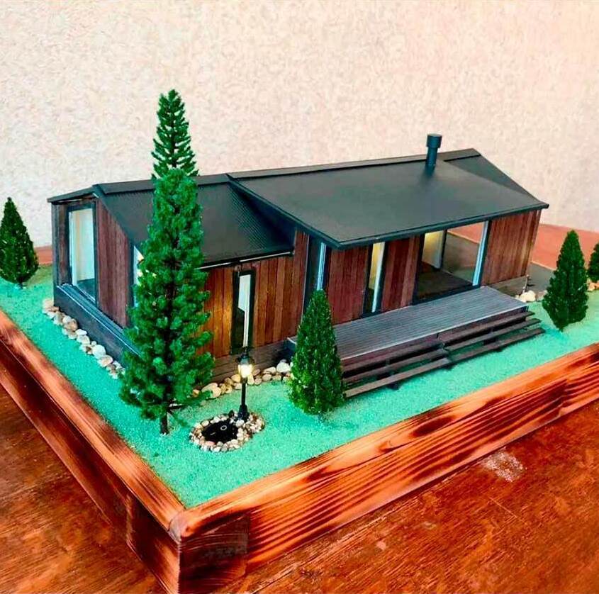  Масштабная модель одноэтажного загородного дома с просторной террасой, ведущей к входной двери по деревянным ступеням. Дом выполнен в современном стиле: фасад облицован деревянными панелями, крыша — тёмная, двускатная, с дымоходом. Большие панорамные окна обеспечивают визуальную лёгкость конструкции. Вокруг дома высажены миниатюрные хвойные деревья, создающие ощущение уединённости и уюта. Придомовая территория оформлена с использованием искусственного газона и декоративных камней. Установлен уличный фонарь в классическом стиле, подчёркивающий эстетику проекта. Макет демонстрирует продуманную планировку участка, сочетание архитектурных и ландшафтных решений, идеально подходит для визуализации проектов частной застройки, презентаций перед заказчиками и использования в экспозициях архитектурных бюро.