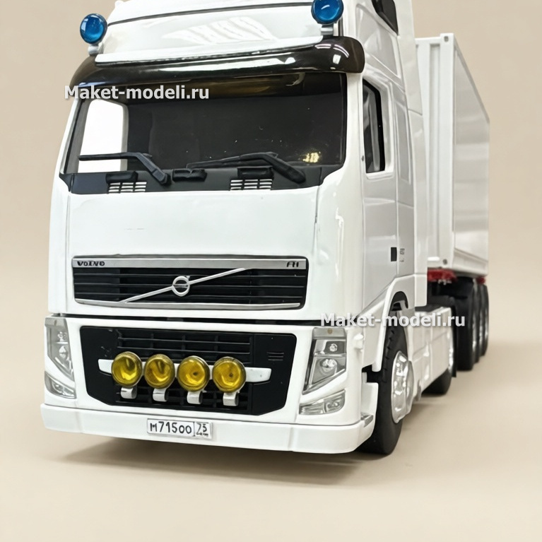 Масштабная модель белого грузового тягача Volvo FH с прицепом изготовлена для подарка руководителю Белая модель грузового тягача Volvo FH с жёлтыми дополнительными фарами и полуприцепом на фоне стеллажа с пластиковыми ящиками