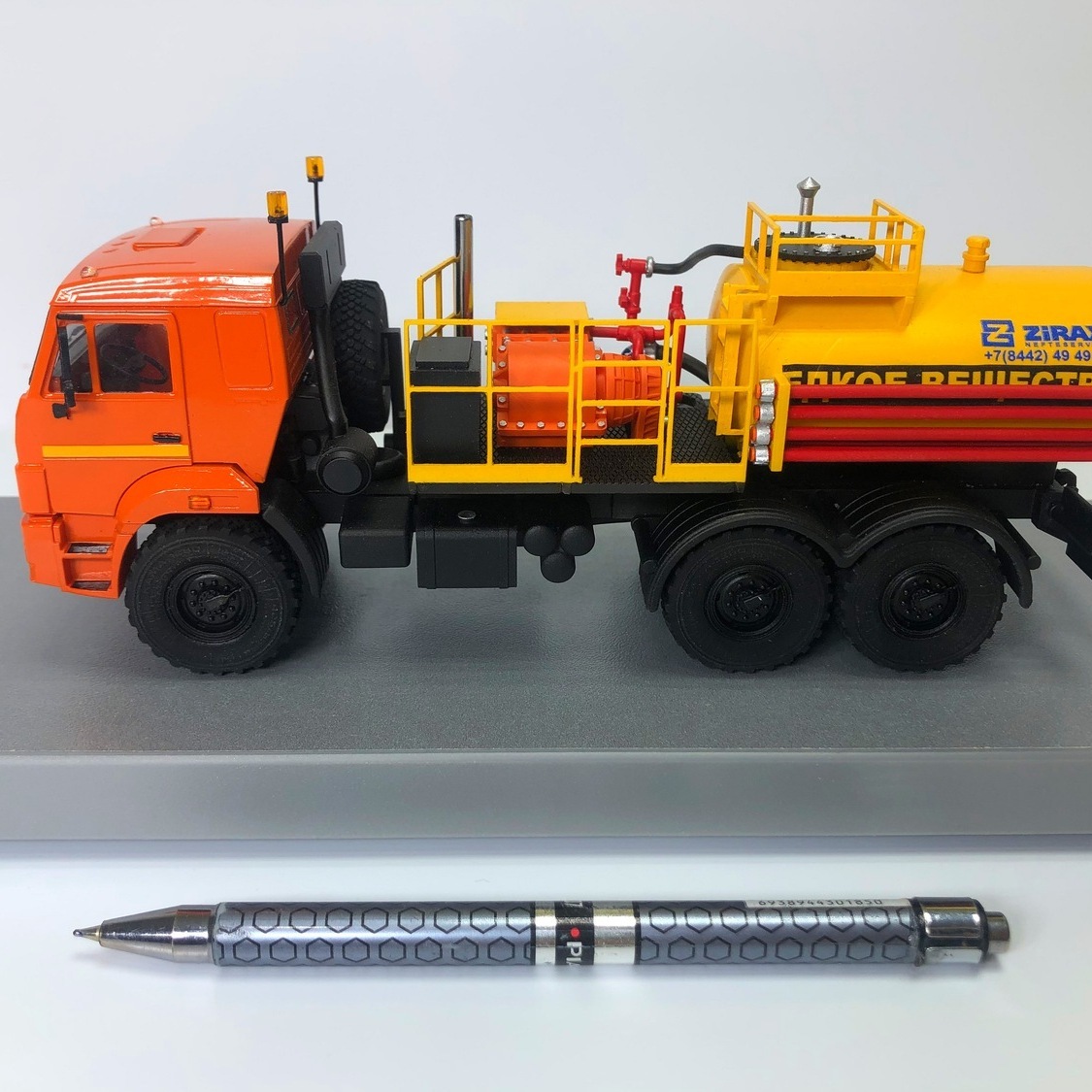 Масштабная модель автоцистерны ZIRAX для жидких веществ – макет спецтехники на подставке изготовленный в макетной мастерской maket-modeli.ru Масштабный макет оранжевого грузовика‑автоцистерны ZIRAX для жидких веществ, вид сбоку, модель установлена на серой подставке, на переднем плане лежит ручка для сравнения размеров