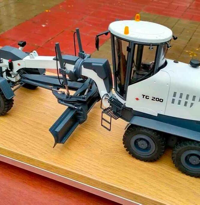 Детализированная масштабная модель автогрейдера TG 200: реалистичный макет дорожно-строительной техники с кабиной, фронтальным отвалом и системой гидравлических рычагов. Модель выполнена в бело-серой цветовой гамме, оснащена имитацией рабочих фар и лестницей для доступа в кабину. Видны крупные колёса с выраженным протектором, элементы ходовой части и конструктивные детали корпуса. Установлена на деревянной подставке, идеально подходит для демонстрации на выставках, использования в презентациях или пополнения коллекции спецтехники.