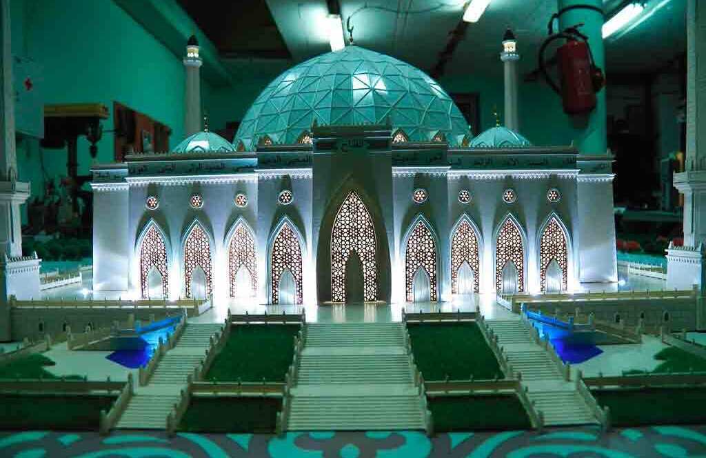Детальный масштабный макет мечети, выполненный с высокой степенью проработки: в центре композиции — массивный геодезический купол, по углам расположены четыре высоких минарета. Фасад украшен изящными арочными проёмами с ажурными решётками, подсвеченными изнутри, что создаёт эффект свечения. Перед мечетью — широкие симметричные ступени, ведущие к главному входу. По обе стороны от ступеней — декоративные бассейны с синей подсветкой, окружённые зелёными газонами. Территория вокруг мечети оформлена с учётом традиционных элементов ландшафтного дизайна: миниатюрные деревья, аккуратные дорожки, орнаментальная плитка. Модель идеально подходит для презентации архитектурных проектов, выставочных экспозиций и образовательных целей, позволяя в деталях рассмотреть особенности исламской архитектуры.
