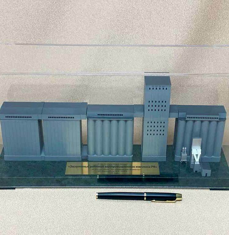 Однотонный серо‑графитовый макет промышленного объекта. • Компоновка типична для элеваторного комплекса: слева два прямоугольных блока, далее секция цилиндрических силосов, в центре высокая норийная (технологическая) башня, справа ещё ряд силосов и небольшие вспомогательные узлы — приемно‑погрузочные и транспортирующие устройства. • Основание тёмное, с металлической табличкой; перед витриной лежит ручка, показывая настольный масштаб модели. • По стилистике это подарочный/представительский макет объекта хранения и перегрузки зерна.