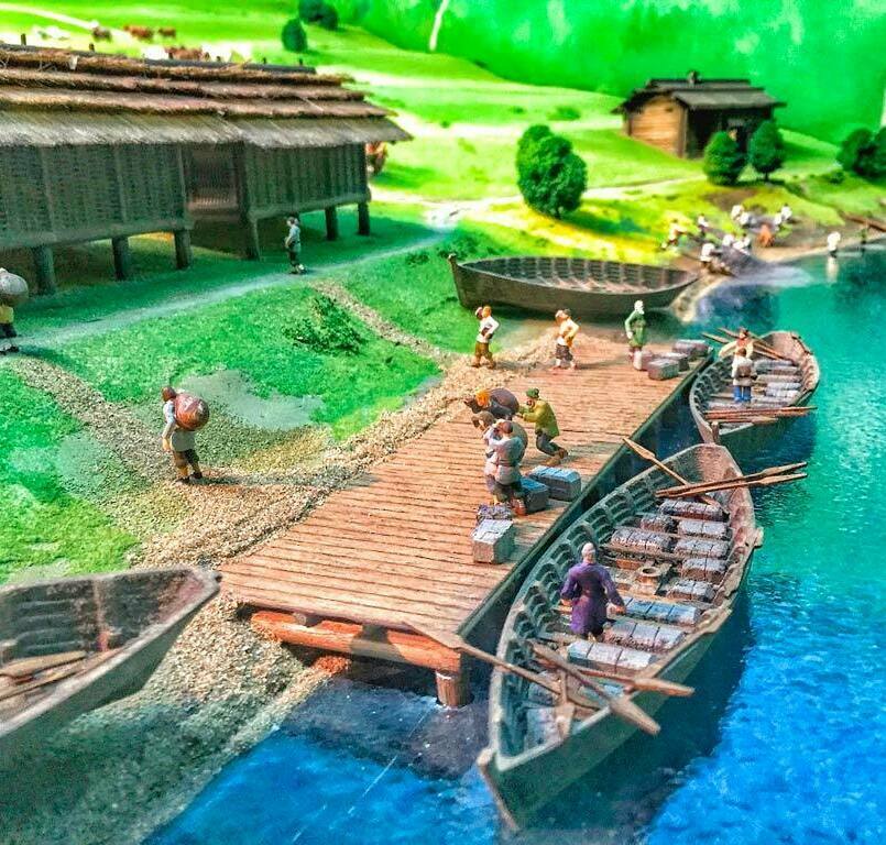 Макет изготовленный для музея на заказ На макете изображена небольшая прибрежная слобода у прозрачной синей реки. К деревянному настилу пристани подошли узкие лодки с вёслами; люди выгружают из них тяжёлые тюки и серые каменные блоки, сгибаясь под ношей и перекликаясь друг с другом. По утоптанной дорожке к реке спускаются новые путники с заплечными мешками. Сбоку возвышается длинное строение на сваях под соломенной крышей — то ли амбар, то ли хозяйственный сарай, где хранятся припасы. Дальше, на зелёном холме, виден отдельный дом с дымоходом и аккуратными деревцами вокруг; у берега суетится ещё группа людей. В глубине макета угадываются пастбища с маленькими фигурками скота. Всё вместе создаёт образ спокойной деревенской жизни у старинного речного тракта, где вода служит дорогой, а пристань — сердцем поселения.