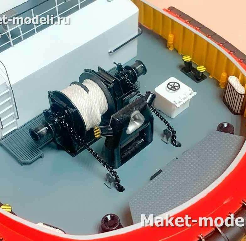 Это детализированный макет узла «буксирное устройство на носовой палубе буксира» с лебёдкой, степлом и швартовной арматурой. • Рабочая палуба буксира с установленной одно­барабанной буксирной лебёдкой (hawser winch). На барабан уложен синтетический буксирный трос; на торце лебёдки — швартовочный шпиль/варпинг‑хед для выборки конца. • Перед лебёдкой — буксирный степл (towing staple, направляющая скоба) для безопасного изменения угла ведения буксирного троса. • Палубная арматура: кнехты (двухрогие бочки), направляющие/клюзы, палубный ящик/пост управления лебёдкой, люк, трап и леерное ограждение. • По периметру фальшборта — мощная резиновая фендровая защита, характерная для портовых буксиров.