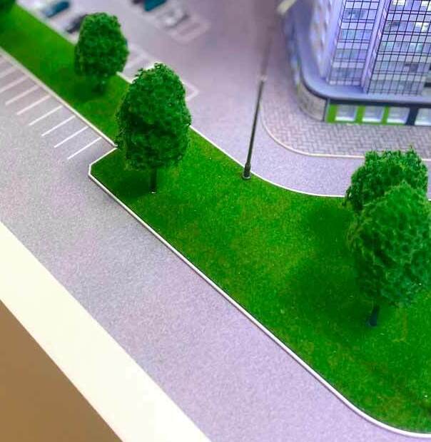 Детализированный макет многоэтажного жилого комплекса с подсветкой: крупные стеклянные фасады, огражденные внутренние дворы, тротуары и парковочные места, зеленые газоны с декоративными деревьями, уличные фонари и элементы благоустройства для демонстрации инфраструктуры и планировки проекта