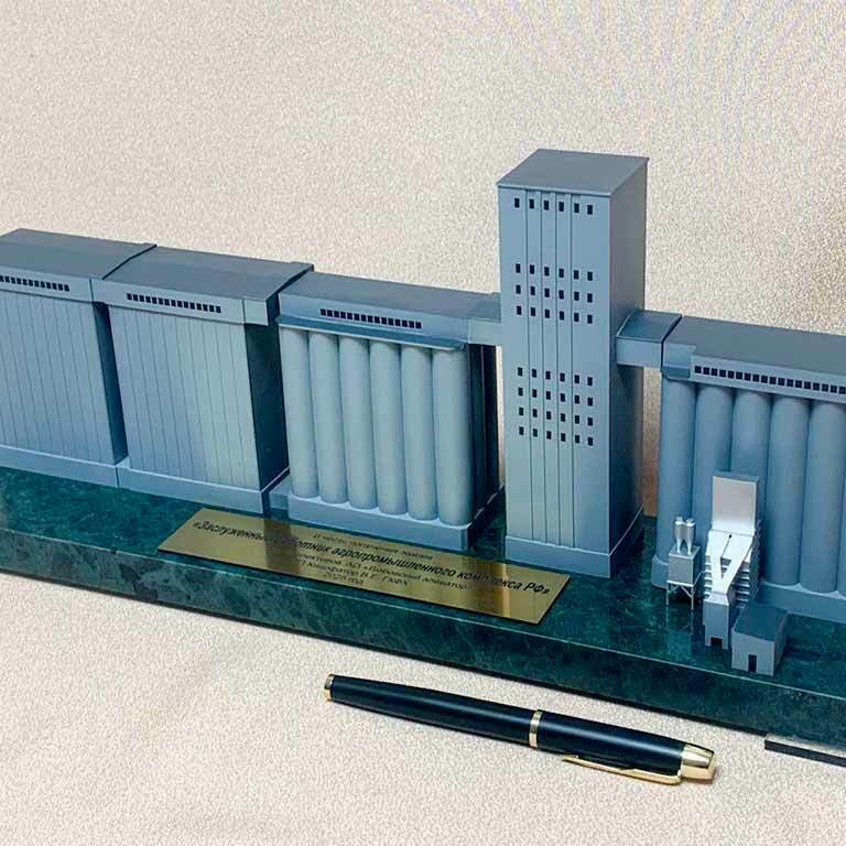 3D-масштабный макет зернового элеватора, демонстрирующий ключевые элементы технологического комплекса: три прямоугольных блока модульных силосов для хранения зерна, центральная группа вертикальных цилиндров (силосные батареи) под общей крышей-галереей, высокая прямоугольная норийно-распределительная башня с технологическими площадками. Справа расположен миниатюрный приёмно-очистительный узел с загрузочной эстакадой и аспирационными фильтрами. Макет установлен на презентационном постаменте с табличкой, рядом лежит ручка для демонстрации масштаба. Детализированная модель наглядно иллюстрирует процессы приёма, очистки, хранения и отгрузки зерна, подходит для использования в выставочных и образовательных целях.