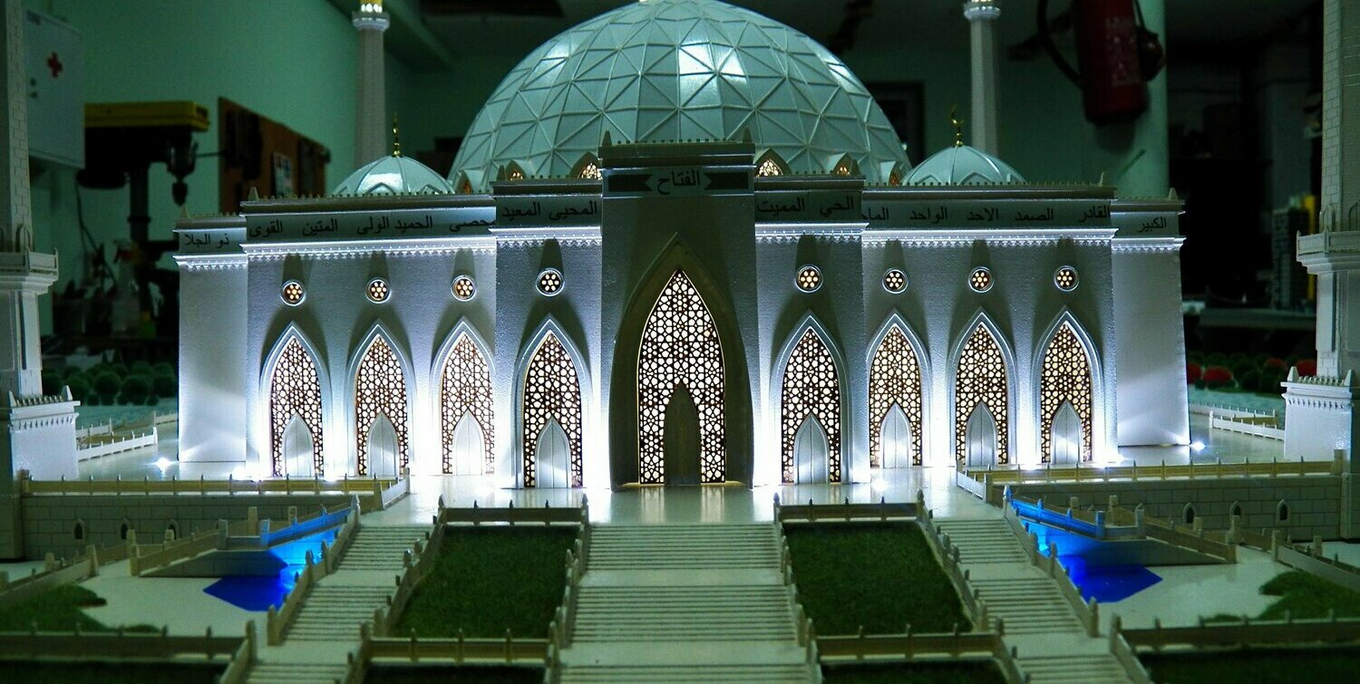 Масштабный настольный макет мечети: белый фасад с резными ажурными арочными воротами и орнаментальными решётками, большой геодезический купол, видимые минареты, фриз с арабской каллиграфией, внутренняя и наружная подсветка, широкие ступени, зелёные газоны и декоративные бассейны с синей подсветкой