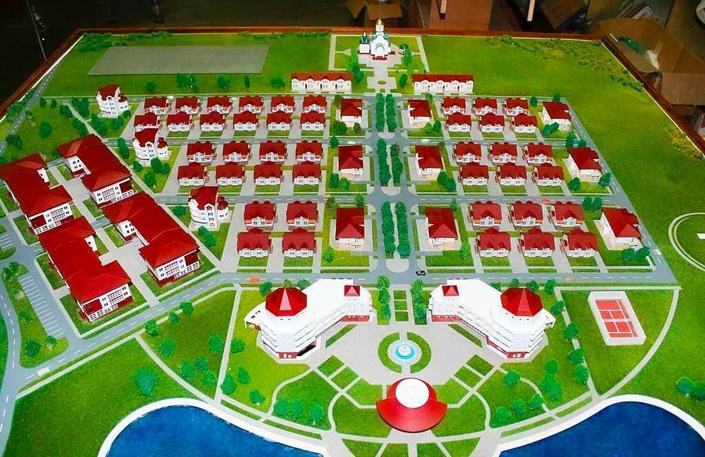 Детальный макет коттеджного посёлка в масштабе: аккуратно выстроенные ряды двухэтажных домов с выразительными красными крышами и белыми фасадами, окружённые миниатюрными деревьями и зелёными газонами. В центре композиции — крупный комплекс с куполами и террасами, напоминающий культурно-развлекательный центр или клубный дом. Справа от центра расположена спортивная площадка с разметкой для игр. В нижней части макета — искусственный водоём с голубой поверхностью, имитирующей воду, и прогулочными дорожками вдоль берега. Видны асфальтированные дороги, тротуары, парковочные зоны и элементы благоустройства: фонари, скамейки, клумбы. Макет демонстрирует продуманную планировку с чётким зонированием: жилая застройка, рекреационная зона, инфраструктурные объекты. Идеален для презентации проектов загородной недвижимости, планирования коттеджной застройки и архитектурных презентаций.