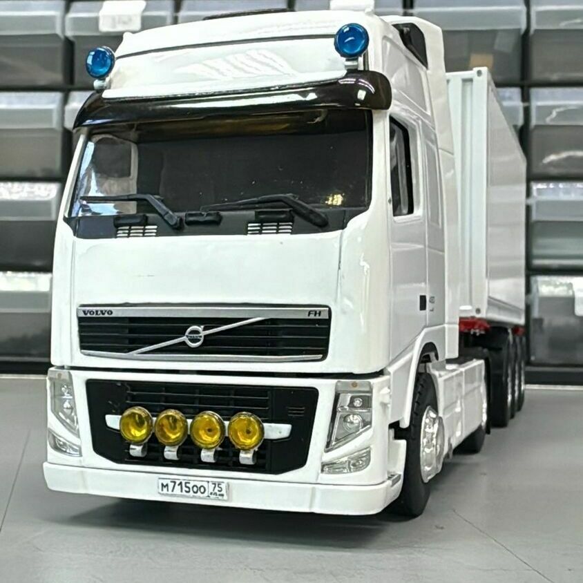 Масштабная модель белого грузового тягача Volvo FH с прицепом изготовлена для подарка руководителю Белая модель грузового тягача Volvo FH с жёлтыми дополнительными фарами и полуприцепом на фоне стеллажа с пластиковыми ящиками