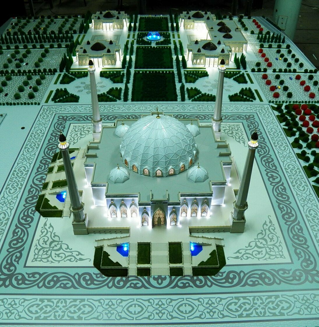 Детализированный макет мечети с геодезическим центральным куполом и четырьмя высокими минаретами, подсвеченные арочные фасады, орнаментальная плитка, террасированные площади, синие декоративные фонтаны и миниатюрные деревья — архитектурная модель для презентации проекта