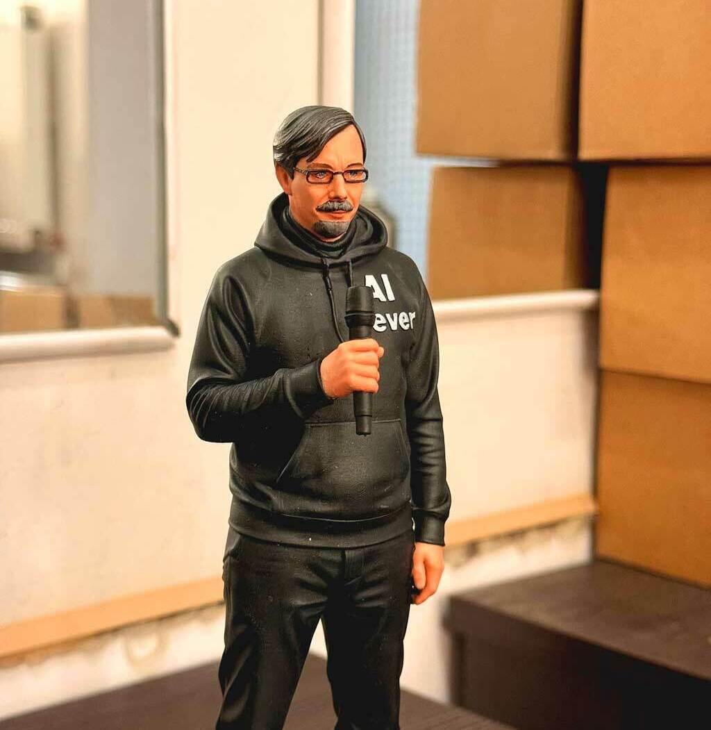 Реалистичная 3D-фигурка человека на заказ по фото: детализированная статуэтка с микрофоном, напечатанная на 3D-принтере. Кастомная модель в чёрной худи с надписью — идеальный сувенир для коллекционеров и поклонников публичных выступлений! 3D-фигурка мужчины, изготовленная по фотографии на 3D-принтере. Статуэтка изображает человека в расслабленной позе с микрофоном в руке. Одет в чёрную худи с надписью «AI ever», чёрные брюки, носит очки и аккуратную бороду. Высокая детализация: проработаны черты лица, текстура одежды, поза передаёт атмосферу публичного выступления. Фигурка отлично подойдёт для украшения офиса, в качестве оригинального подарка или элемента коллекции 3D-моделей. Пример персонализированной печати с сохранением индивидуальных особенностей.