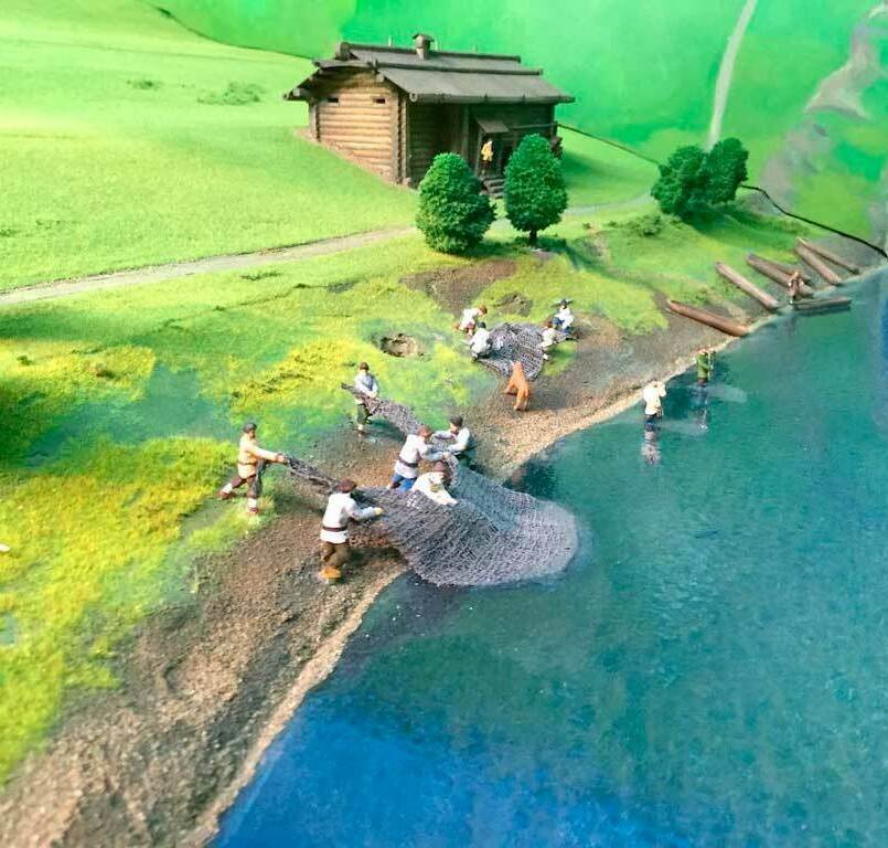 Макет изготовленный для музея на заказ подробный макет речного берега. У прозрачной синей воды, где галька мягко переходит в травянистый откос, несколько фигурок рыбаков тянут тяжёлый невод к суше. Часть людей стоит в воде по колено, другие, упершись ногами в мокрый песок, помогают вытащить сеть. По берегу разбросаны свёрнутые снасти, рядом стоит лошадь, чуть поодаль кто‑то чинит сети. На пригорке, окружённая невысокими деревьями, возвышается деревянная изба с крыльцом — единственное строение среди зелёных холмов. В стороне, у воды, аккуратно сложены брёвна или узкие лодки, готовые к спуску на реку.