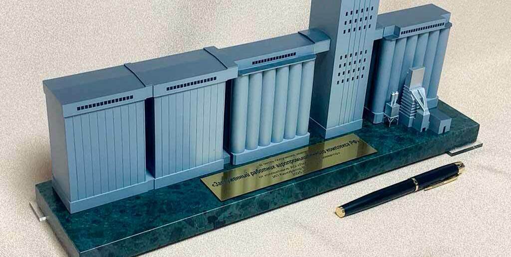Линейно выстроенный комплекс зданий: два прямоугольных корпуса (склады/приёмные секции), блок вертикальных цилиндрических силосов и высокая рабочая башня посередине. • Справа — вспомогательные узлы: площадка со ступенями/лестницей и небольшие технические блоки. • Все элементы выполнены в едином серо‑голубом цвете, с прорисованными вентиляционными решётками и окнами. • Макет установлен на полированной подставке «под камень» с металлической табличкой с надписью на русском; рядом лежит ручка для масштаба.  Суть изображения: Это презентационный настольный макет промышленного зерноперерабатывающего/зернохранилищного объекта — элеватора с силосами и рабочей башней для приёма, хранения и перегрузки зерна.