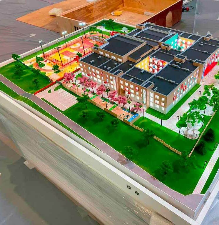 Масштабная модель школьного комплекса: четырёхэтажное здание с выразительным фасадом, большими окнами и функциональными пристройками. На макете детально проработаны внутренние помещения (классы, актовый зал, столовая), видимые через прозрачные секции крыши. Прилегающая территория включает спортивную площадку с разметкой для игр, зону отдыха с беседками, зелёные насаждения, пешеходные дорожки и парковочные места. Реалистичная проработка элементов ландшафта (газоны, деревья, кустарники) и малых архитектурных форм. Макет демонстрирует продуманную планировку образовательного учреждения, сочетающую учебные, спортивные и рекреационные зоны, и идеально подходит для архитектурных выставок, презентаций проектов и образовательных мероприятий.