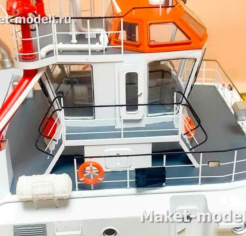  Ходовая рубка (пилот-хаус) с панорамным остеклением и ярко-оранжевой крышей для визуальной заметности. • Крылья мостика с наружными трапами, леерными ограждениями и противоскользящим покрытием палуб. • Противопожарный лафетный ствол (водяная пушка) красного цвета с подводом трубопровода — типичное оснащение буксиров/спасателей. • Спасательные средства: круги на леерах, герметичный контейнер надувного плота. • Водонепроницаемые двери и люки, шкафы и вентиляционные решётки на надстройке. • Швартовые кнехты/тумбы по периметру палубы (частично в кадре).  Назначение узла: • Управление судном и наблюдение при буксировке/маневрах. • Доступ к лафетному стволу и постам наблюдения при противопожарных работах в акватории порта.