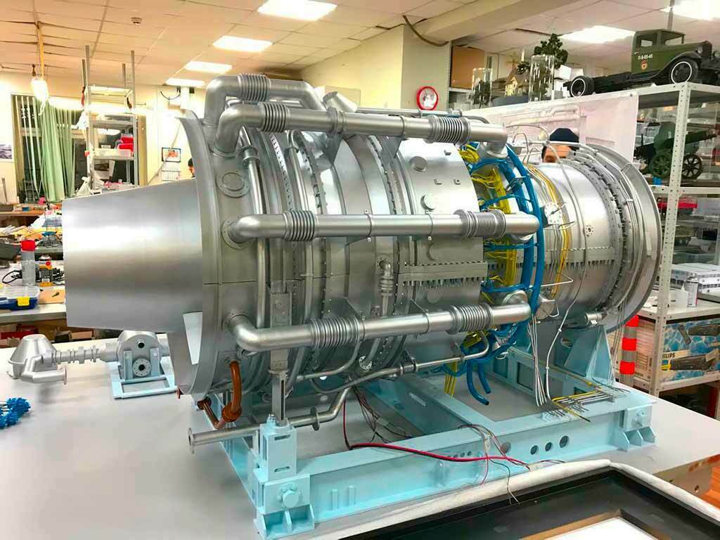 Детализированный макет газовой турбины промышленного оборудования: отчётливо видны лопатки турбинного колеса, корпус с рёбрами жёсткости, разветвлённая система трубопроводов (синие и жёлтые линии), крепёжные элементы, монтажные узлы и элементы системы охлаждения. Модель установлена на демонстрационной раме, демонстрирует внутреннюю компоновку и ключевые узлы газотурбинной установки. Подходит для использования на отраслевых выставках, в образовательных целях, для презентаций инженерных решений в сфере энергетического оборудования. Проработка мелких деталей подчёркивает сложность и точность конструкции промышленного агрегата.