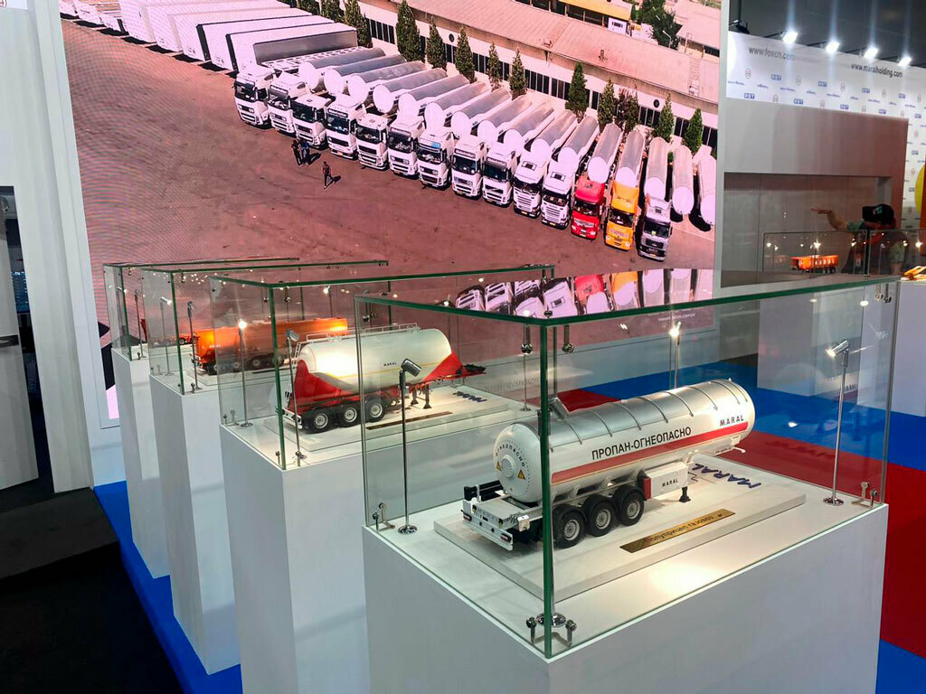 Модель полуприцепа-цементовоза MARAL в стеклянной витрине: белый цилиндрический бак с жёлтой полосой, красная задняя часть, детально проработаны оси, колёса и крепёжные элементы. На баке видна маркировка «MARAL». Экспонат размещён на белой подставке с металлической табличкой, демонстрирует конструкцию современного цементовоза — подходит для выставок, посвящённых грузовому транспорту и спецтехнике, а также для коллекционных экспозиций. Макеты полуприцепов для грузовых автомобилей на выставке в г. Москва