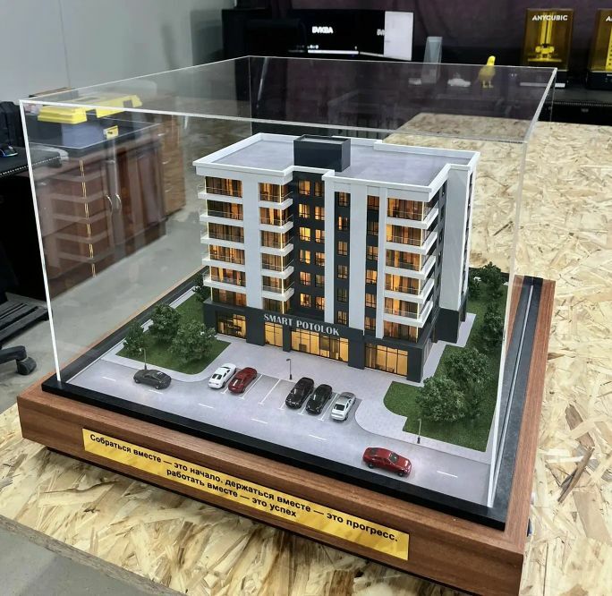 3D-масштабный макет современного многоэтажного жилого здания с подсветкой фасадов. Модель демонстрирует планировку комплекса: главный корпус с балконами и большими окнами, входная группа, крытая парковка с несколькими автомобилями, благоустроенные зелёные зоны с деревьями и кустарниками по периметру. Видны элементы городской инфраструктуры — проезжая часть с разметкой, тротуар, уличные фонари. Макет выполнен с высокой детализацией, включая текстуры фасадов, ландшафт и миниатюрные автомобили, что позволяет наглядно представить будущий объект и его интеграцию в городскую среду. Идеален для презентации девелоперских проектов инвесторам и будущим жильцам.